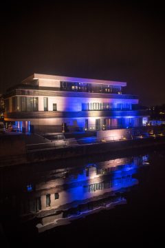 Modern Gebäude mit blauer Beleuchtung, reflektiert im Wasser bei Nacht.